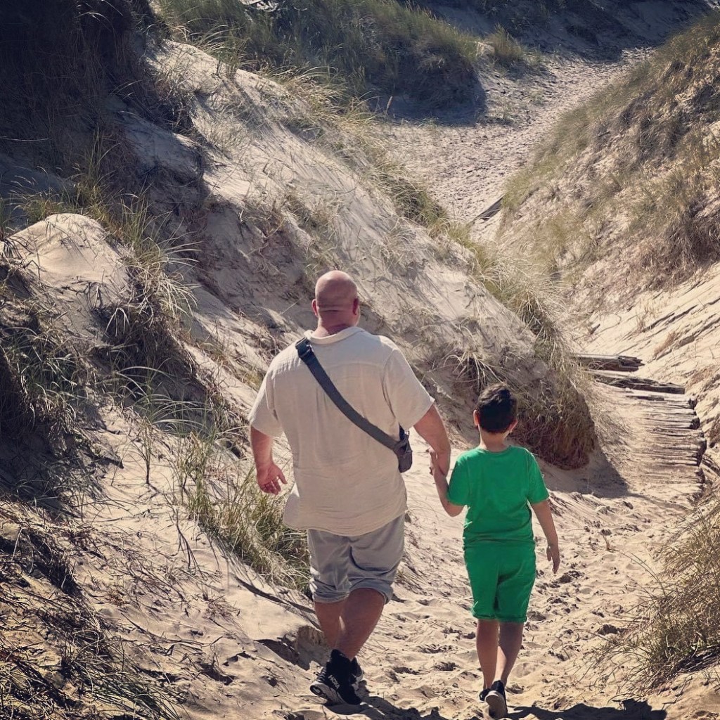 En voksen og et barn går hånd i hånd gjennom sanddyner på en solrik dag.